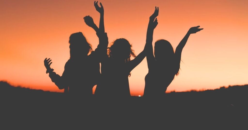 女性3人が手を挙げて楽しそうに踊っている夕暮れ時の写真